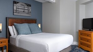A neatly made bed with a wooden headboard, a nightstand with a lamp, a television on a wooden dresser, and a framed picture on the wall. - Getaway on the Riverwalk @ Riverside Suites- 1 Br Condo (San Antonio)