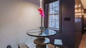 A room with a round glass table, a potted orchid, a window with a view of buildings, and a wooden stool.