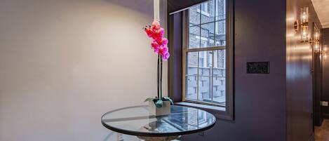 Una habitación con una mesa redonda de vidrio, una orquídea en maceta, una ventana con vista a edificios y un taburete de madera.