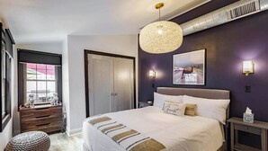 A modern bedroom with a large bed, a hanging light fixture, a framed picture on the wall, and a window with blinds.
