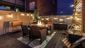 An outdoor patio with a wooden deck, a table with chairs, a fire feature, string lights, and a brick wall in the background.