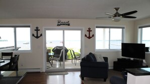 Una sala de estar con televisor, un sofá y un comedor con sillas y una mesa. Hay ventanas con vistas al agua y una puerta que da al exterior.
