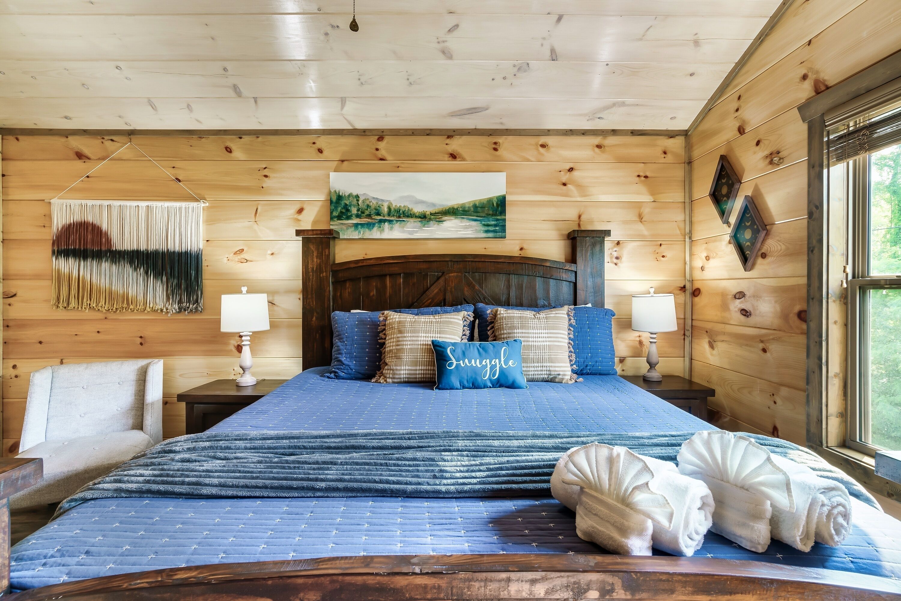 A wooden cabin bedroom with a bed, pillows, a painting, and a window.