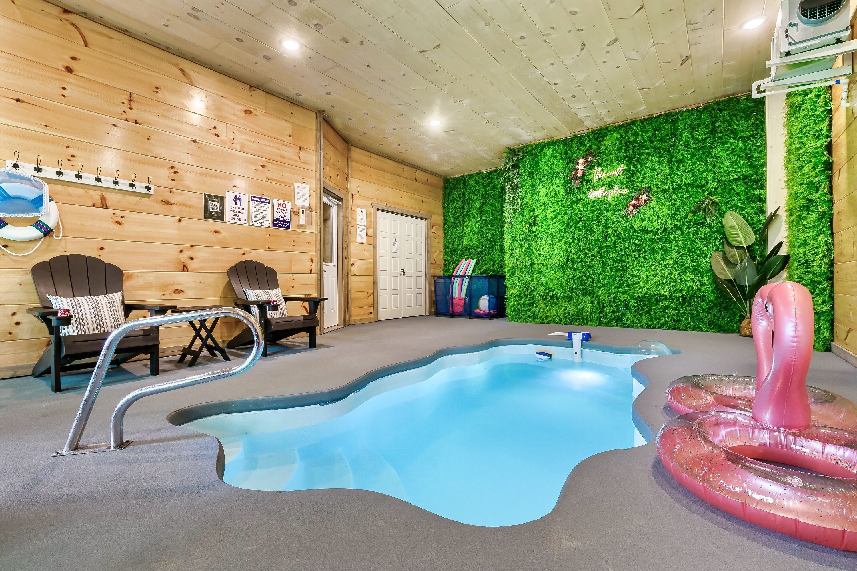 Une piscine intérieure avec un mur en bois et une décoration murale verdoyante, deux chaises et un flotteur gonflable en forme de flamant rose.