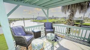 A covered outdoor seating area with wicker furniture and patterned cushions, overlooking a body of water and a grassy landscape.
