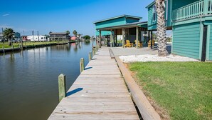 A wooden dock leading to a green building with a covered area and a palm tree.
