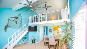 A spacious living area with a high ceiling, a balcony, a dining table, and a TV mounted on the wall.