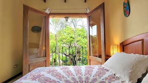 A bedroom with a bed, a nightstand, a window with an open door, and a view of greenery outside.
