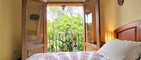 A bedroom with a bed, a nightstand, a window with an open door, and a view of greenery outside.