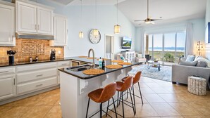 A modern kitchen with white cabinets, a central island with stools, a large window with a view, and a ceiling fan.