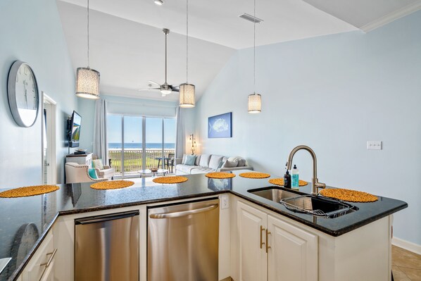 A modern kitchen with a central island, stainless steel appliances, pendant lighting, and a view to the outside. - Top Floor~Beachfront~Heated Pool~Pelican Place (Dauphin Island)