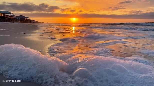 A beach with a sunset, waves, and a pier with buildings.