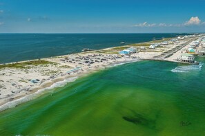 An aerial view of a coastal area with a beach, a pier, and buildings along the shore. - Top Floor~Beachfront~Heated Pool~Pelican Place (Dauphin Island)