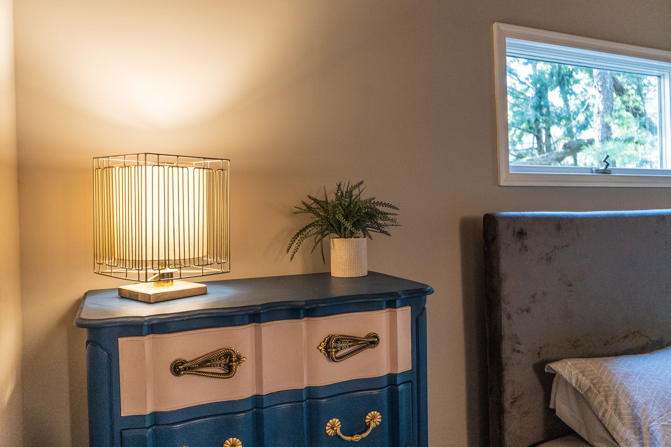 A bedroom with a bedside table, a lamp, a plant, and a window with a view of trees.