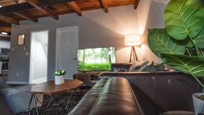 Una sala de estar con un sofá de cuero, una mesita con una planta, una lámpara y un televisor que muestra un paisaje de bosque.