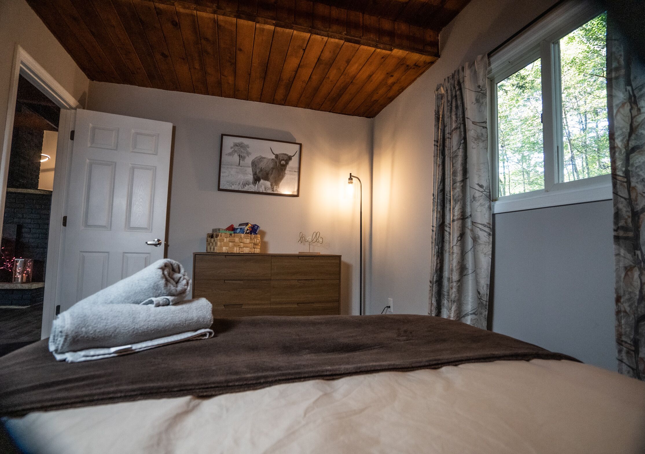 A bedroom with a wooden ceiling, a bed with a brown blanket, a dresser, a framed picture on the wall, and a window with curtains.