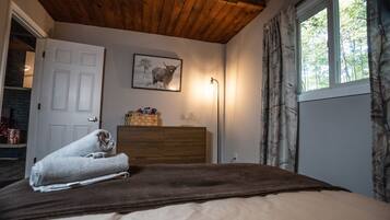 A bedroom with a wooden ceiling, a bed with a brown blanket, a dresser, a framed picture on the wall, and a window with curtains.