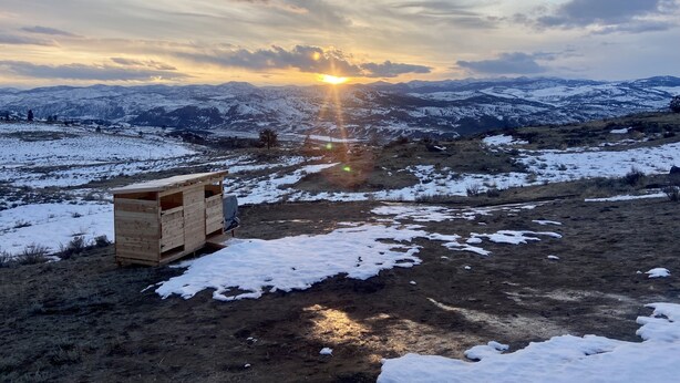 A wooden cabin in a snowy landscape with a view of distant mountains and a sunset.