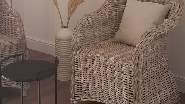A wicker chair with a cushion, a small round table, and a tall vase with pampas grass.