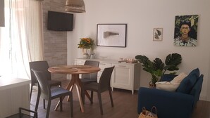 A modern living room with a round wooden table, two chairs, a blue sofa, a large potted plant, and a kitchen area with a white cabinet and a flat-screen TV.