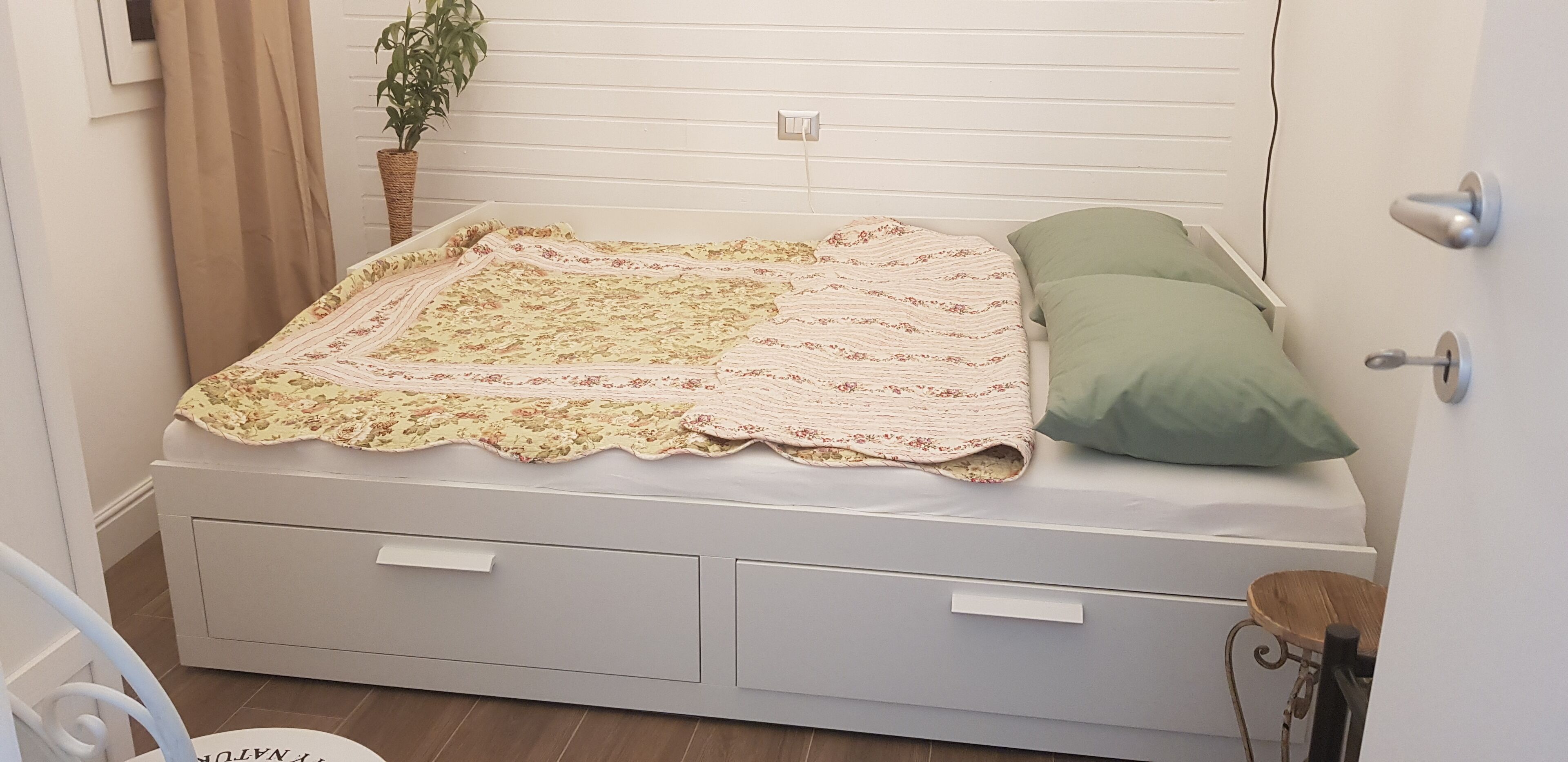 A neatly made bed with a floral bedspread and two pillows, a bedside table with a lamp, and a potted plant on the wall.