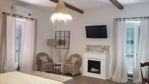 A bedroom with a bed, a fireplace, wicker chairs, a TV, and a ceiling fan.
