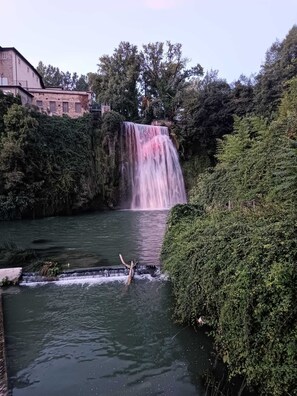Una cascata che si getta in un fiume, circondata da vegetazione lussureggiante e un edificio sullo sfondo.