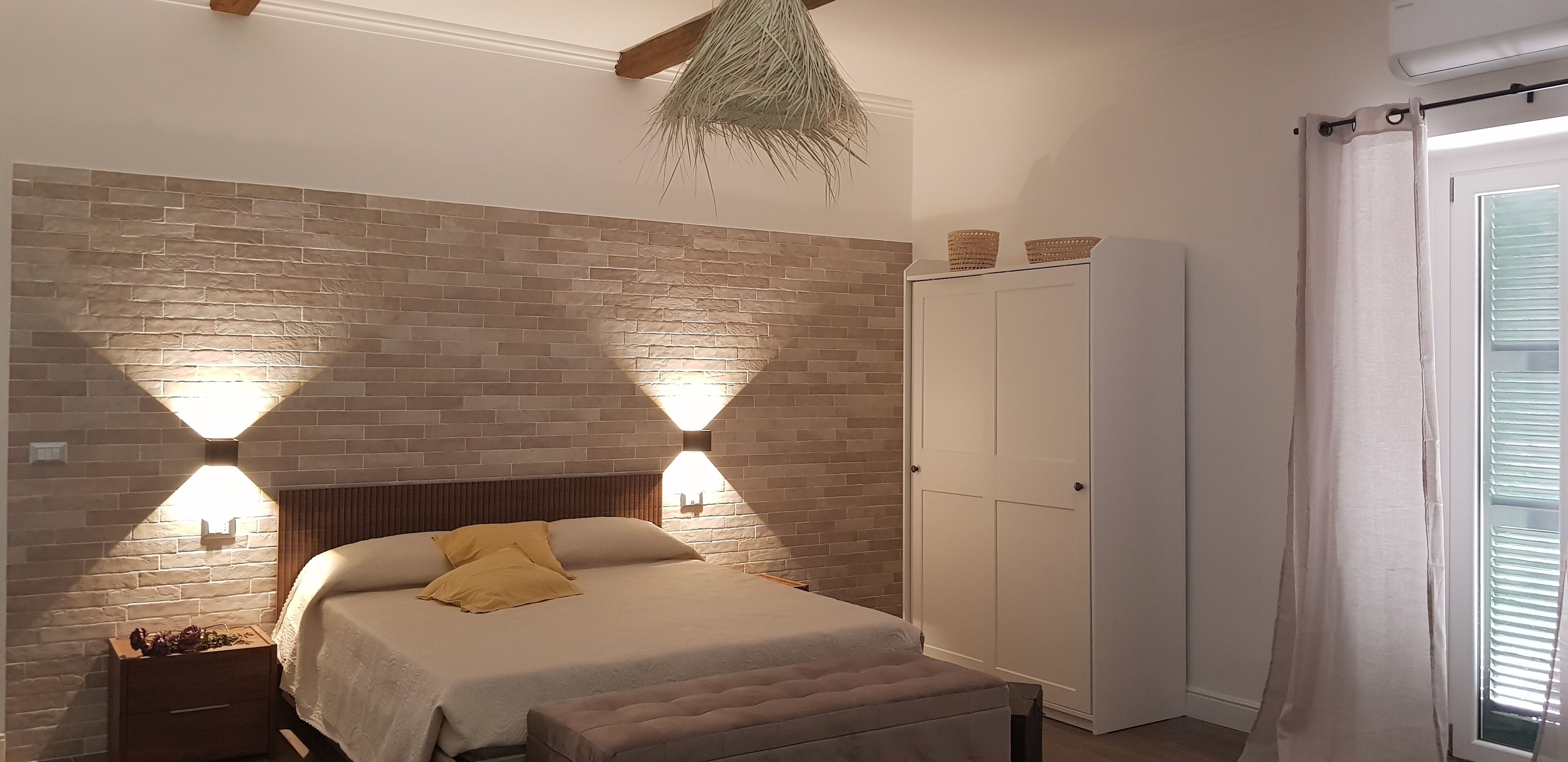 Ein Schlafzimmer mit einer ziegelmusternden Wand, einem Bett mit Kopfteil, zwei Wandleuchten, einem Kleiderschrank und einem Fenster mit Vorhängen.