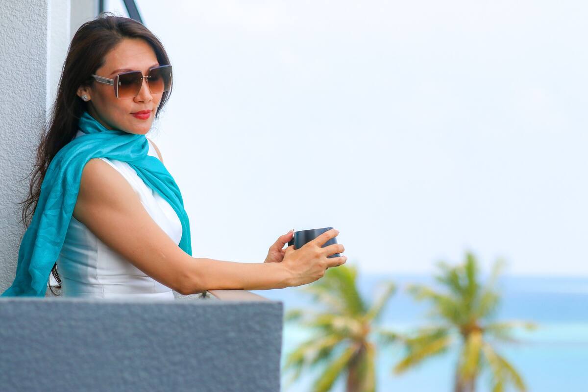 A person holding a mug, leaning on a balcony with a view of palm trees and the sea.