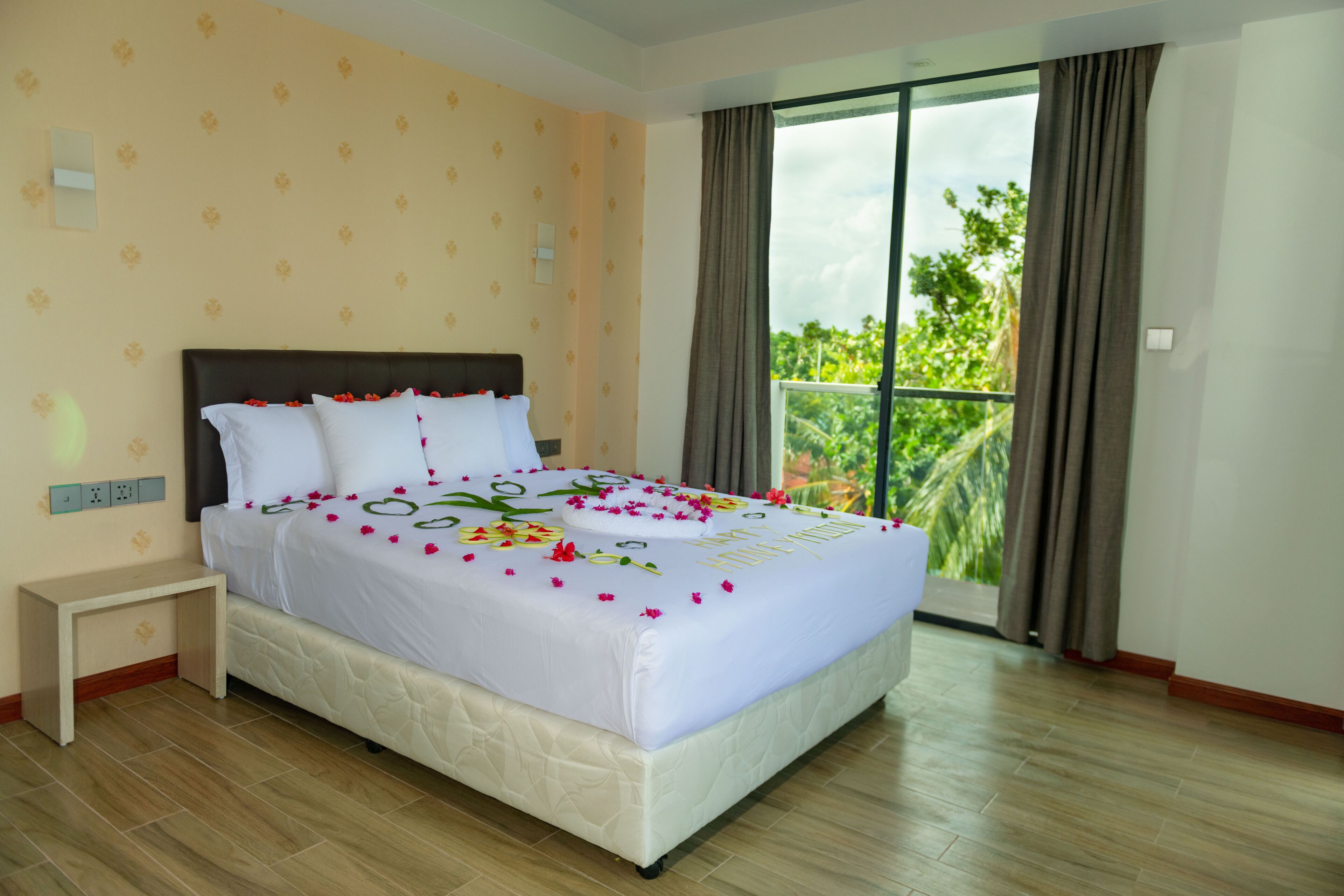 A hotel room with a bed, a nightstand, a window with curtains, and a view of greenery outside.