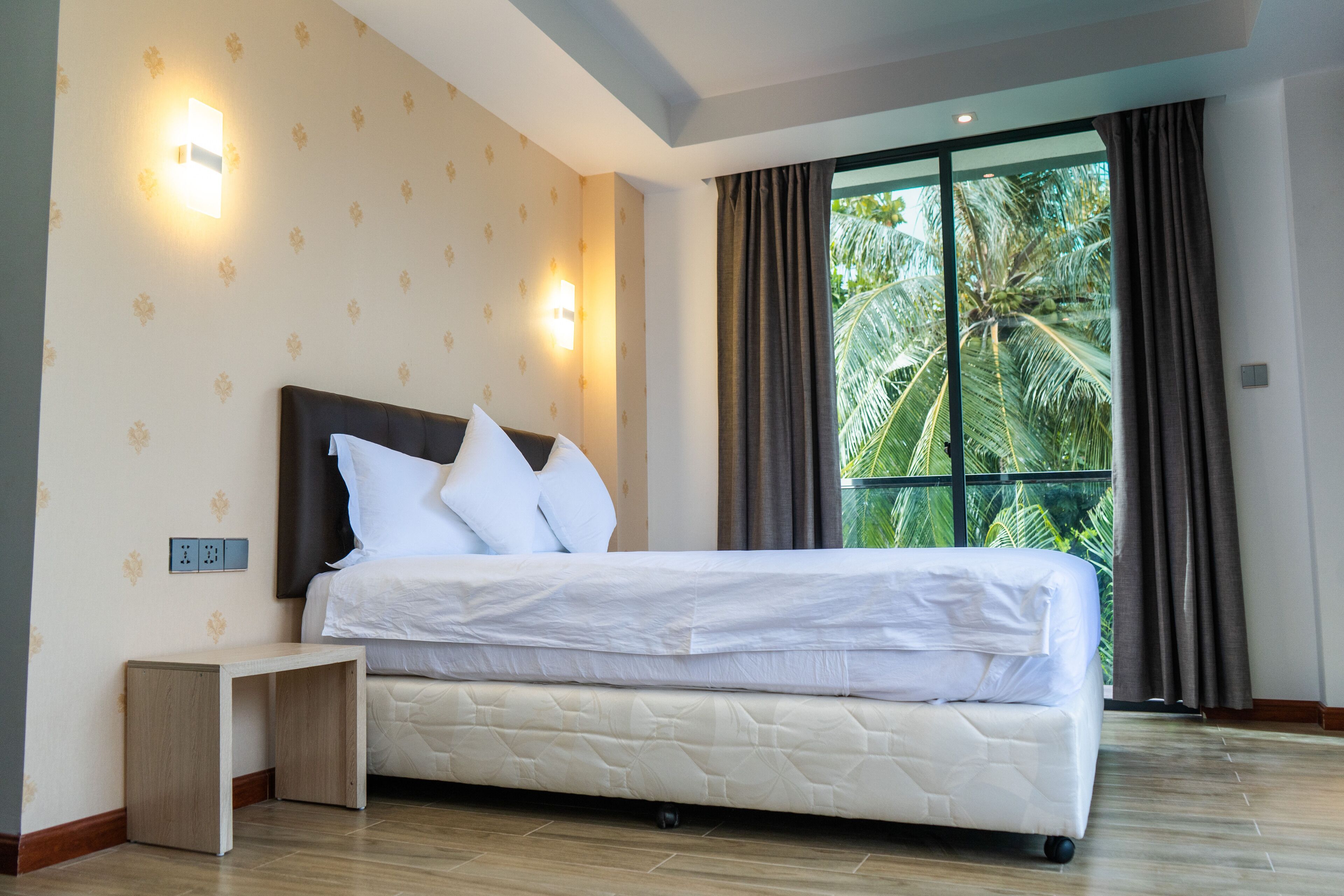A modern hotel room with a large bed, two wall-mounted lights, a small bedside table, and a view of palm trees through a large window.