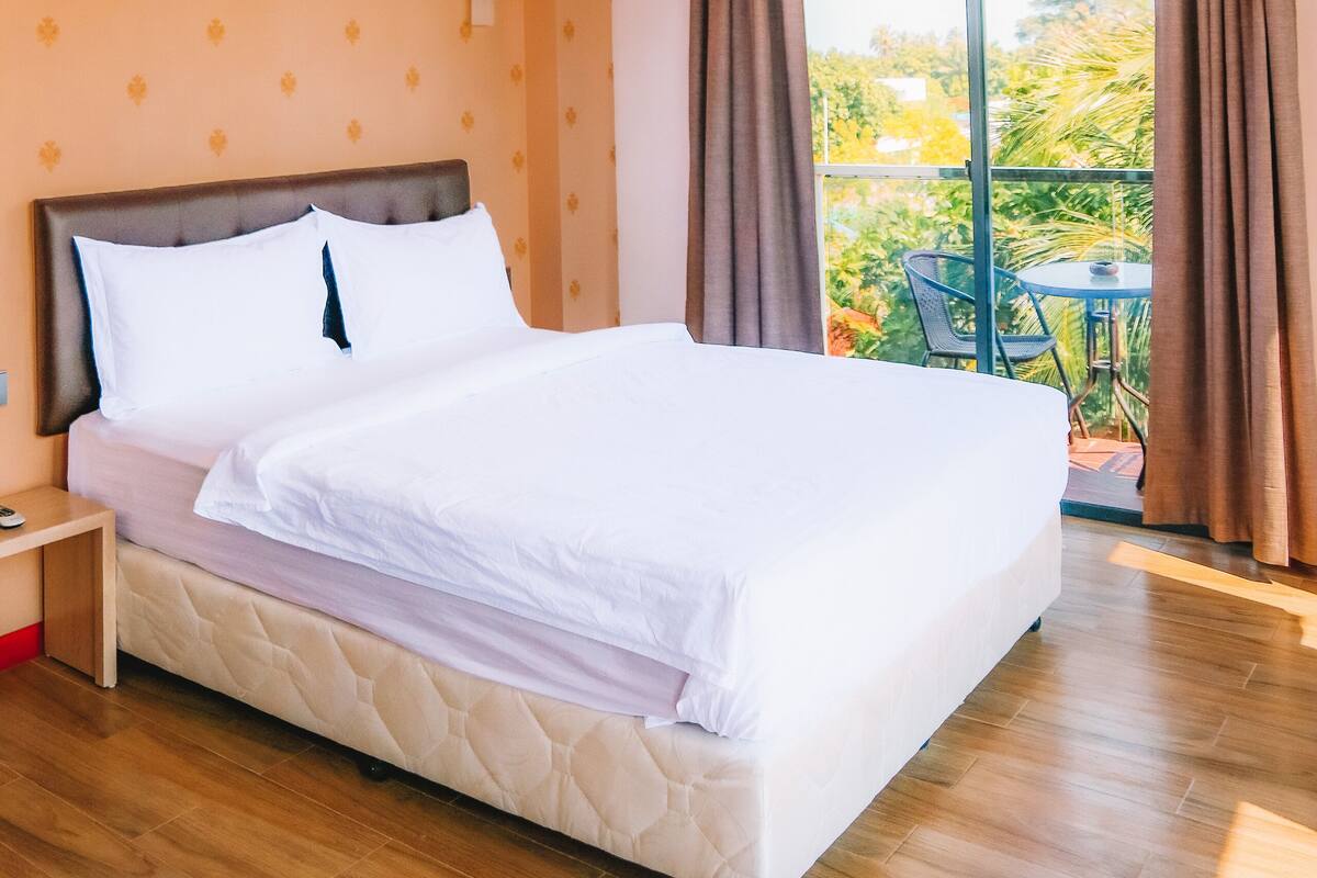 A hotel room with a large bed, a nightstand, and a view of a balcony with a table and chairs.