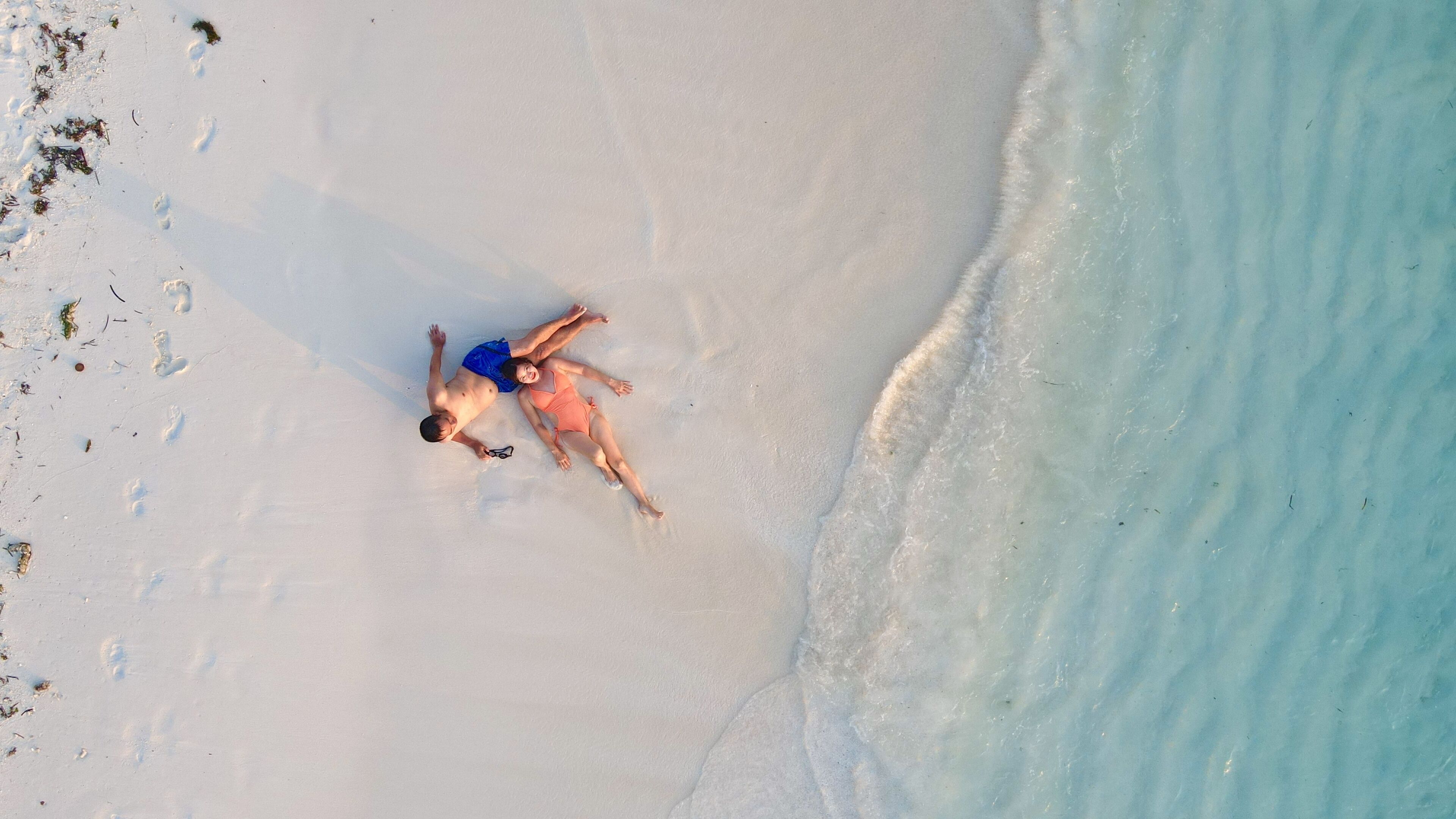 Luftaufnahme eines Strandes mit zwei Personen, die im Sand liegen, bewölktem Himmel und sanften Wellen, die an die Küste rollen.