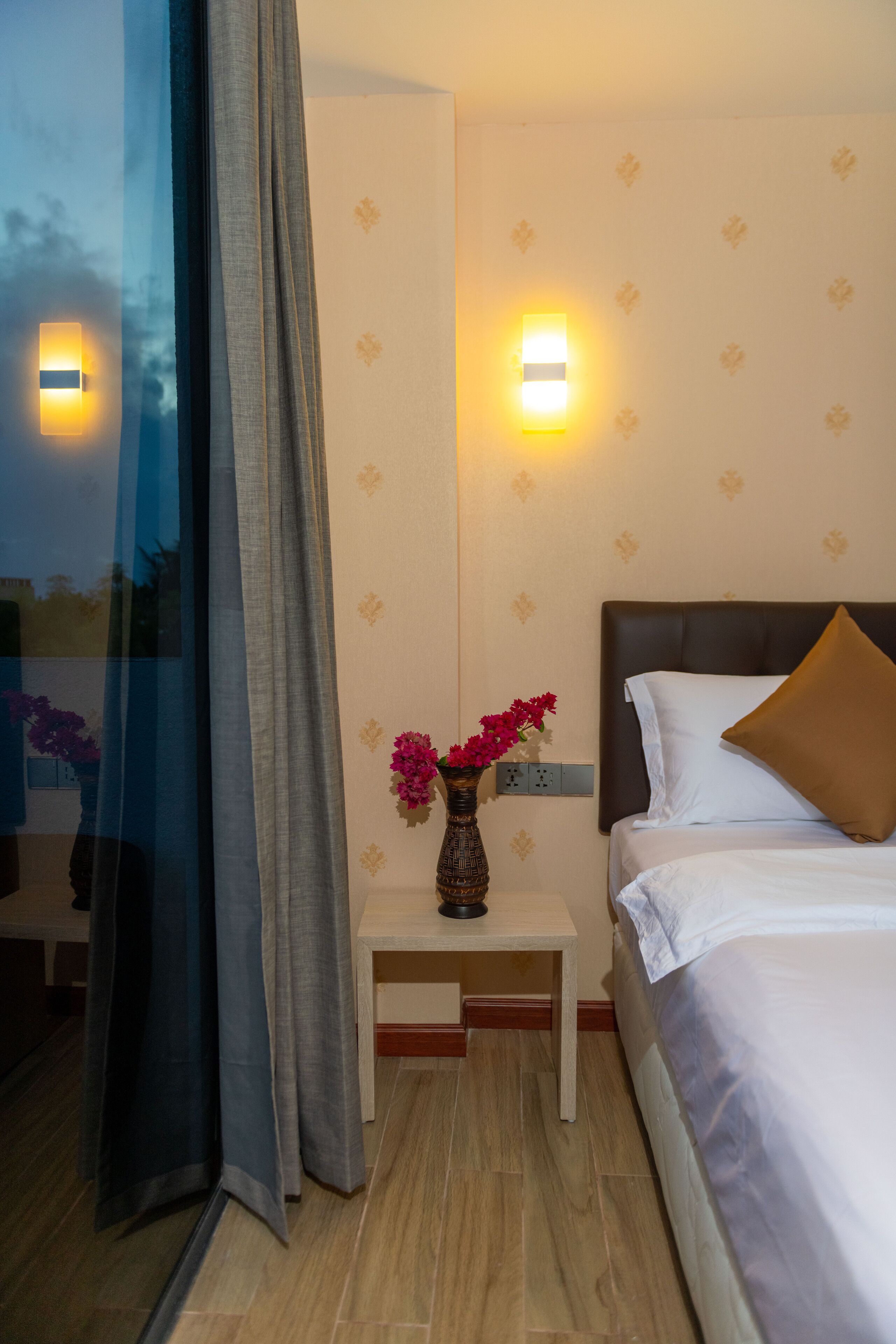 Quarto de hotel com uma cama, mesa de cabeceira com um vaso de flores, luminária e arandela.