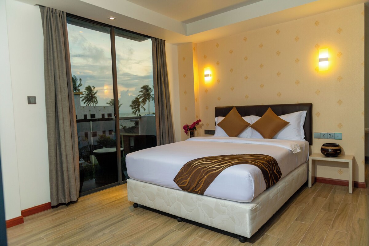 Una habitación de hotel con una cama grande, una mesita de noche con un florero y vistas a palmeras a través de una puerta corrediza.