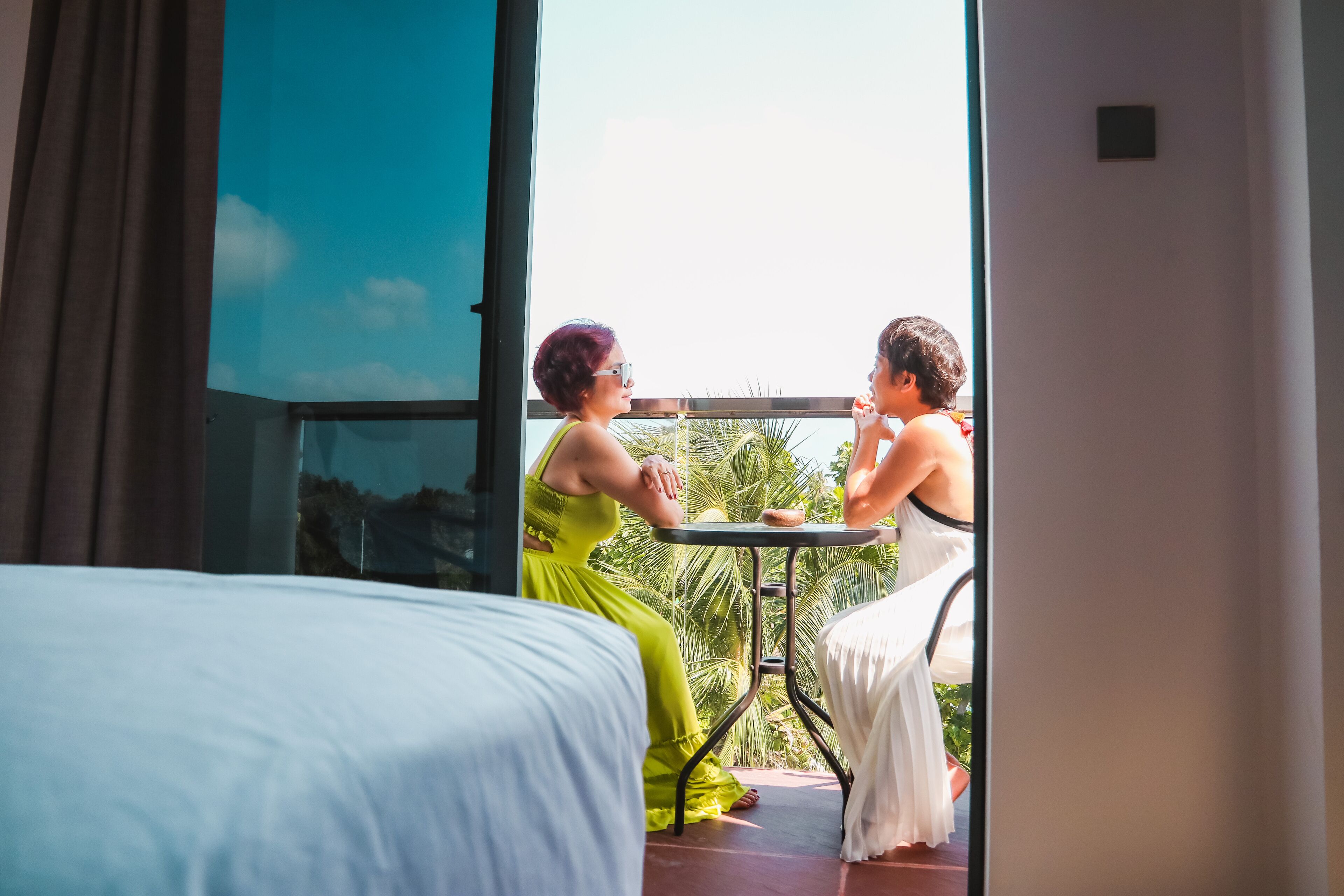 A room with a bed, a balcony with a table, and a view of the sky.