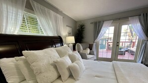 Ein Hotelzimmer mit einem großen Bett, weißer Bettwäsche, einem hölzernen Kopfteil, einem Sessel, einer Topfpflanze und Blick auf die Terrassenanlage.