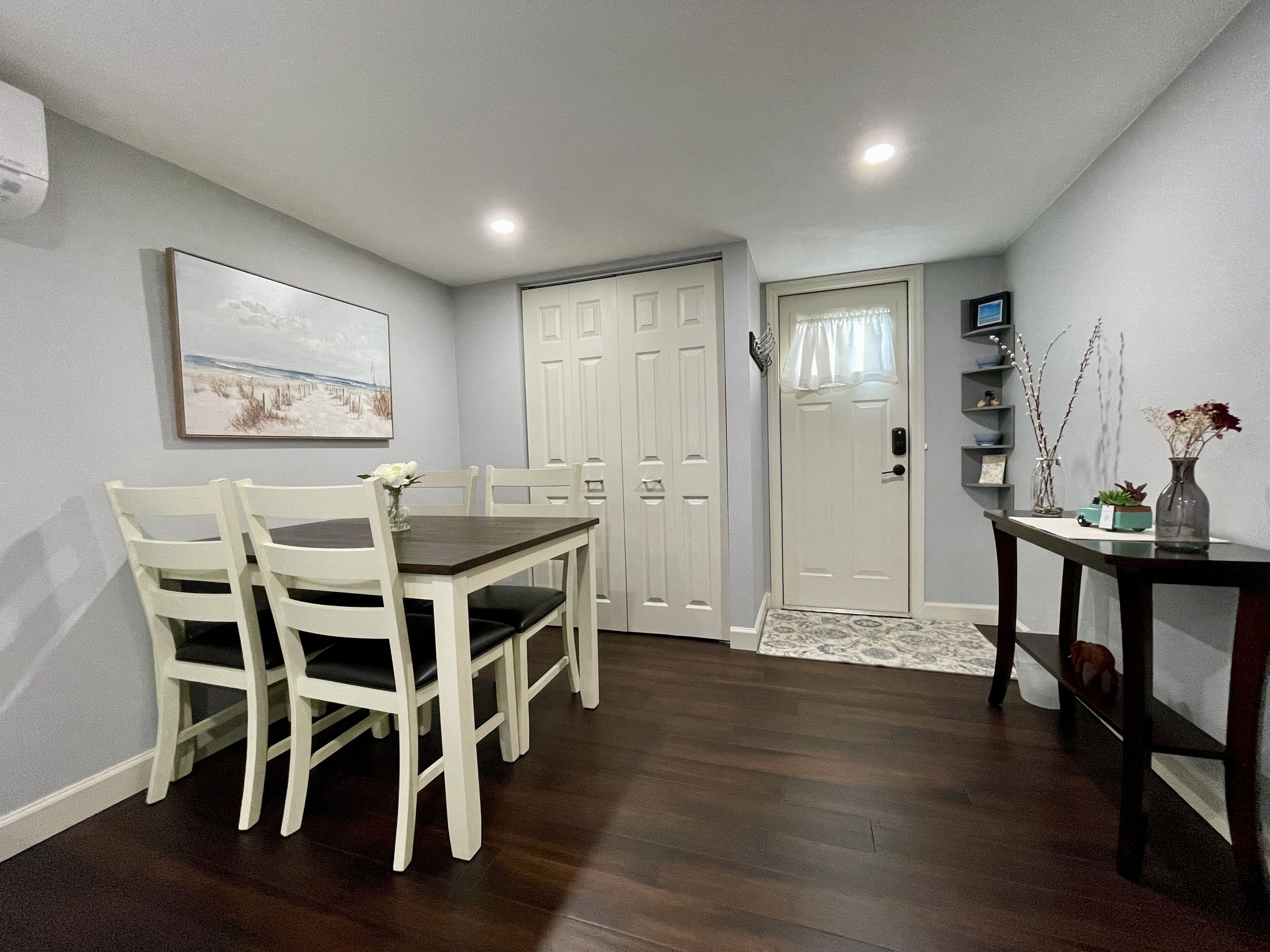 A dining area with a wooden table and white chairs, a white door, and a small side table with a vase.