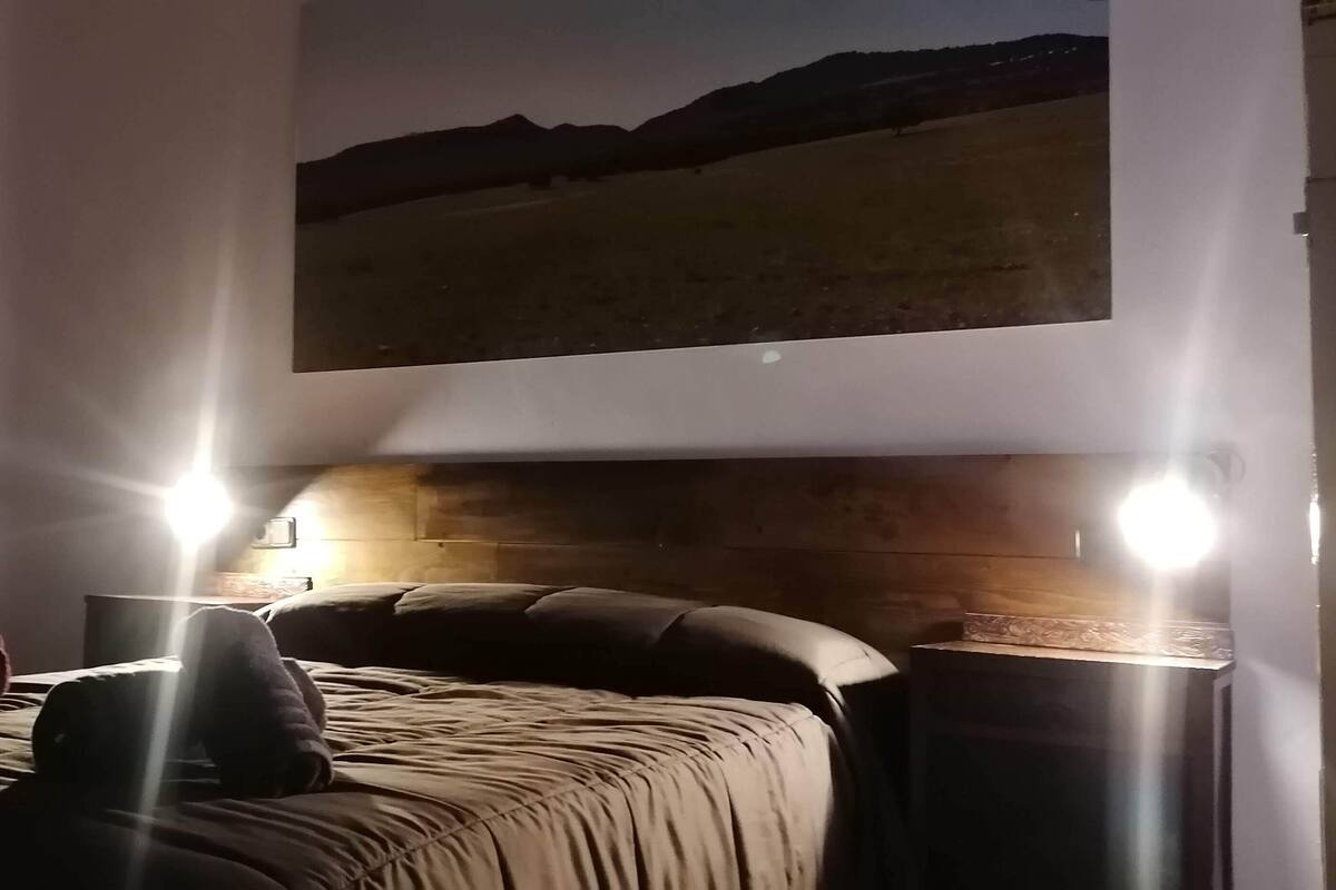 A bedroom with a bed, a nightstand, a lamp, a painting on the wall, and a window with curtains.