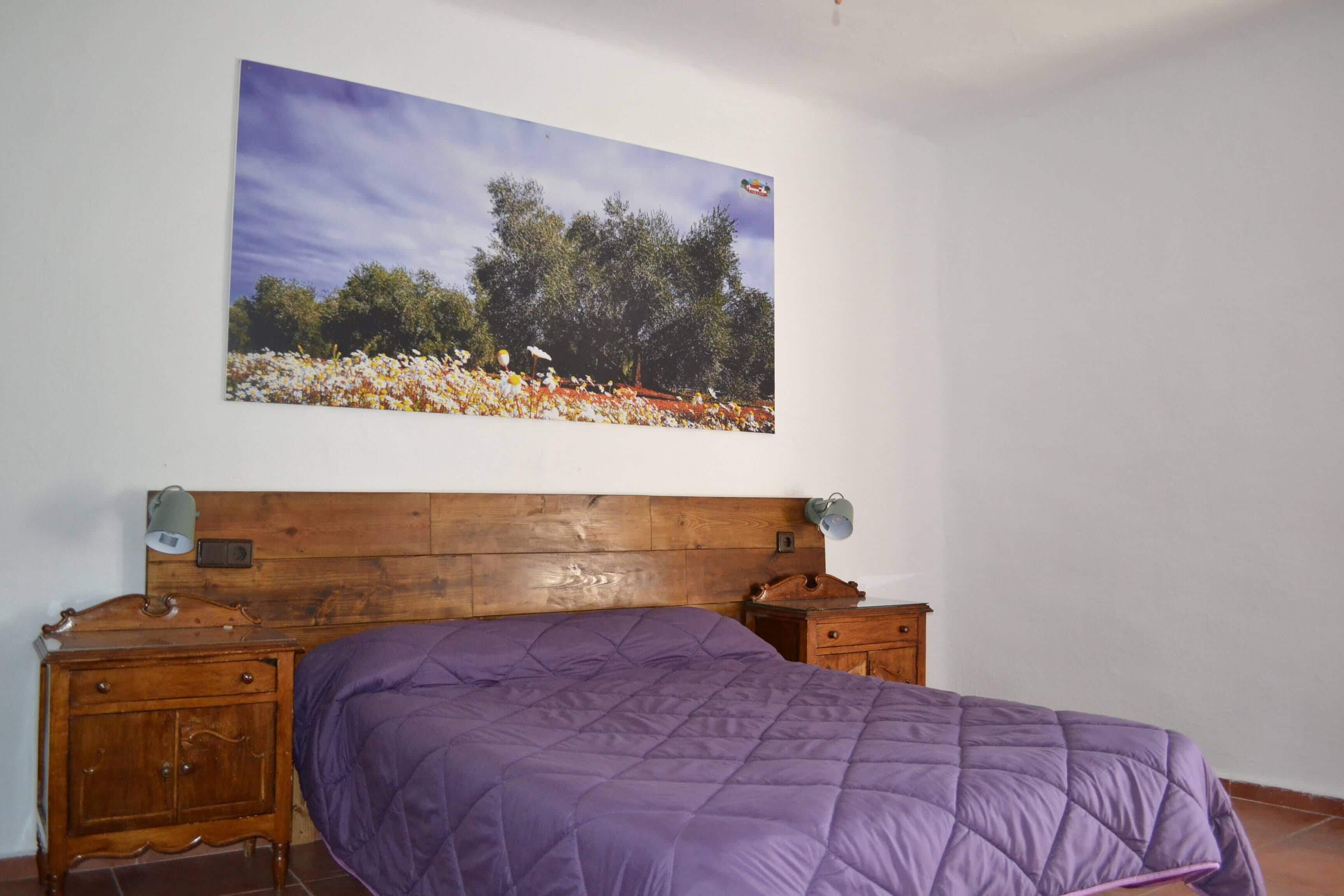 Une chambre à coucher avec une tête de lit en bois, une couvre-lit violette, une table de chevet avec une lampe et un tableau accroché au mur représentant un champ avec des arbres.