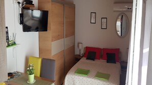 Quarto com cama, escrivaninha, cadeiras, televisão e espelho.
