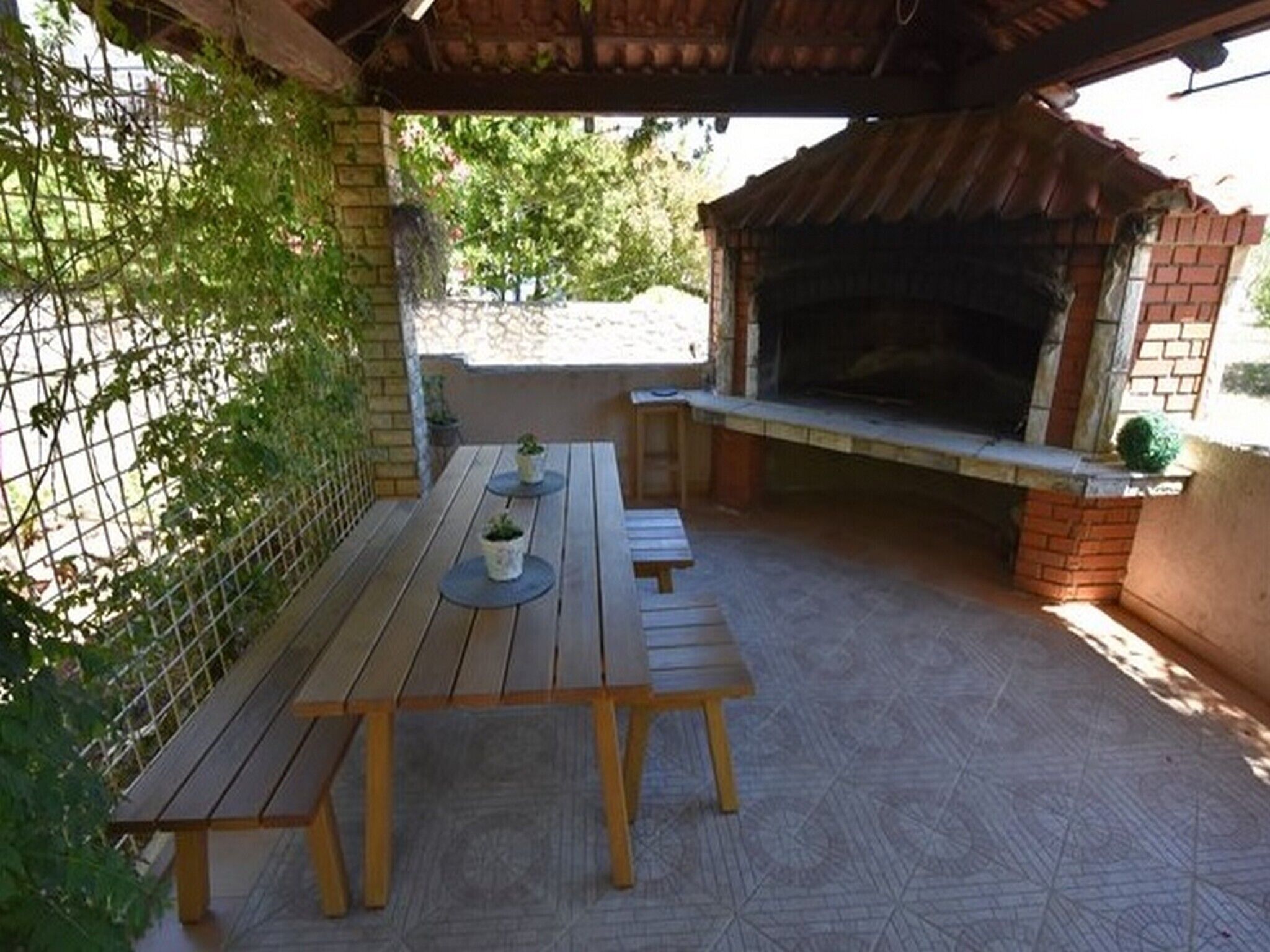 Une terrasse extérieure avec une table et des bancs en bois, un four en briques et une clôture en métal.