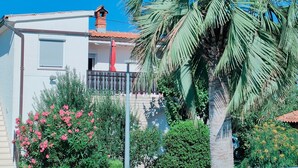 Une maison à deux étages dotée d’un balcon, entourée d’une végétation luxuriante et d’un palmier.