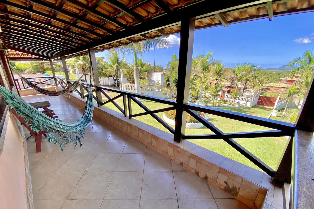 A covered outdoor area with a hammock, tiled floor, and a view of a green landscape with palm trees.