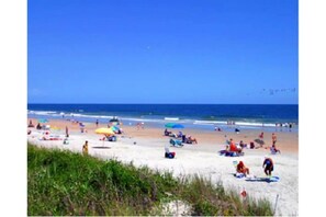 Ein Strand mit badenden Menschen, Sonnenschirmen und einem wolkenlosen, blauen Himmel.