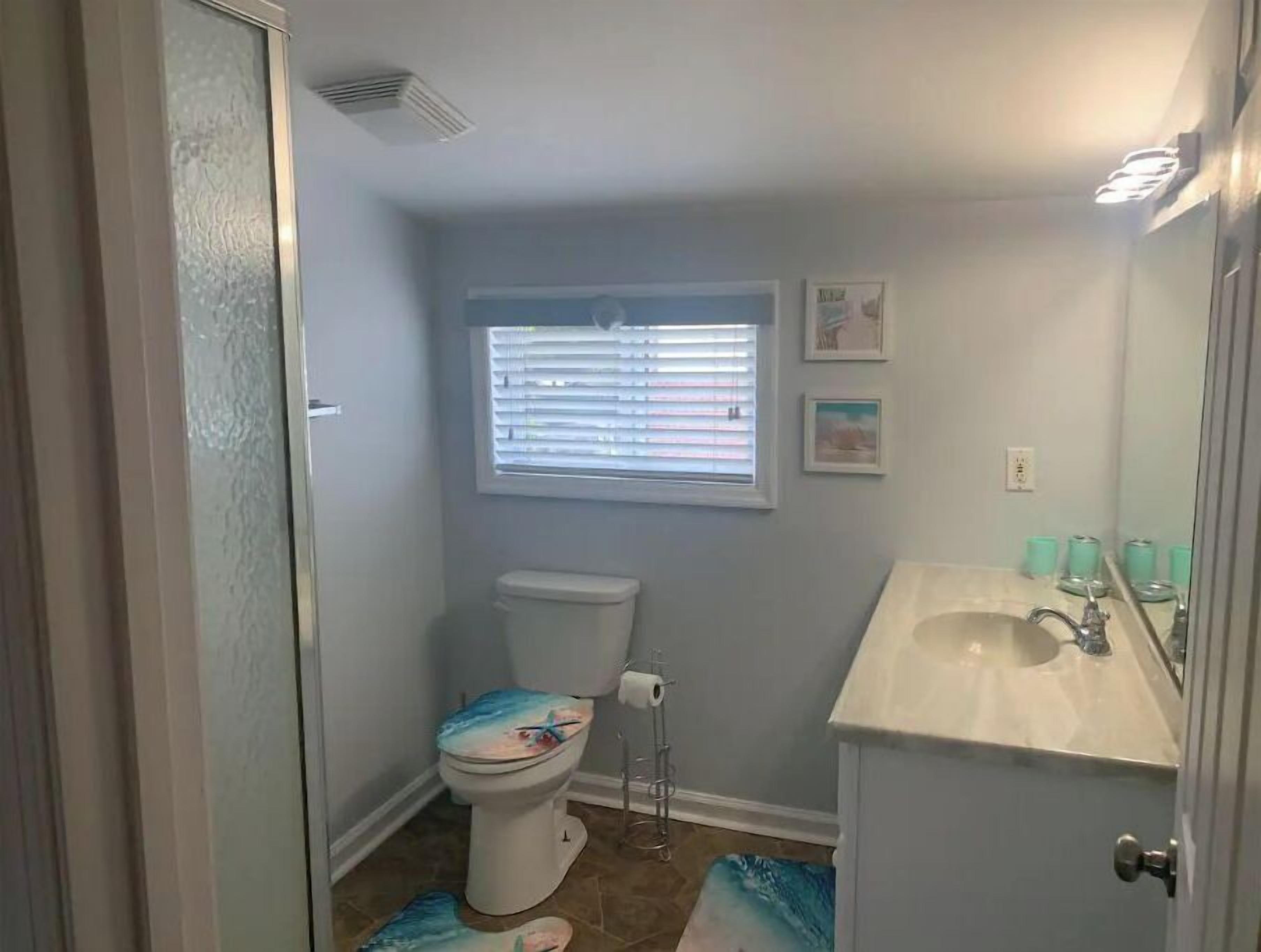 A bathroom with a toilet, a window with blinds, a sink, and a mirror.