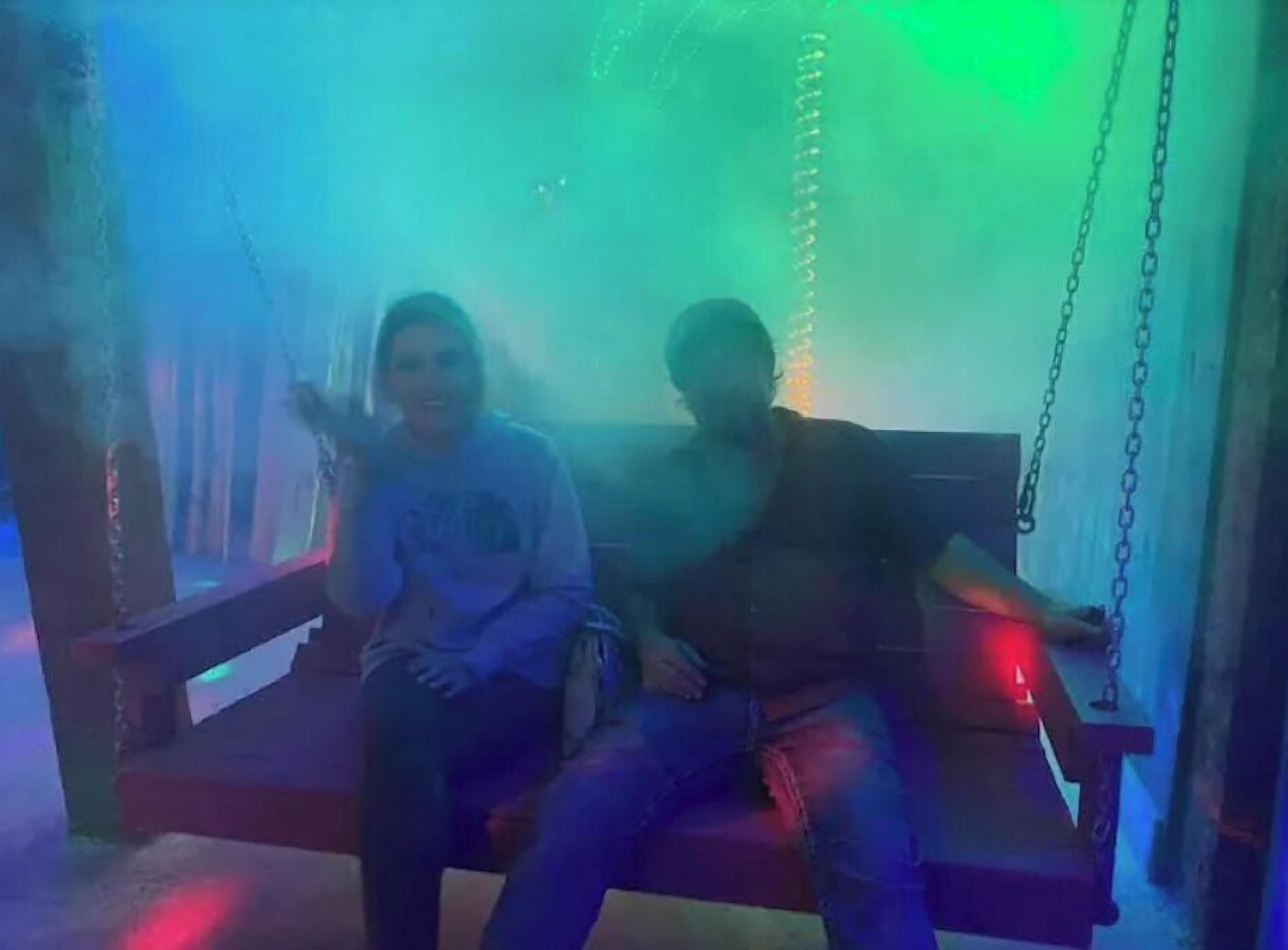 Two people sitting on a swing with chains, in a dimly lit room with blue and green lighting.