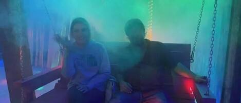 Two people sitting on a swing with chains, in a dimly lit room with blue and green lighting.