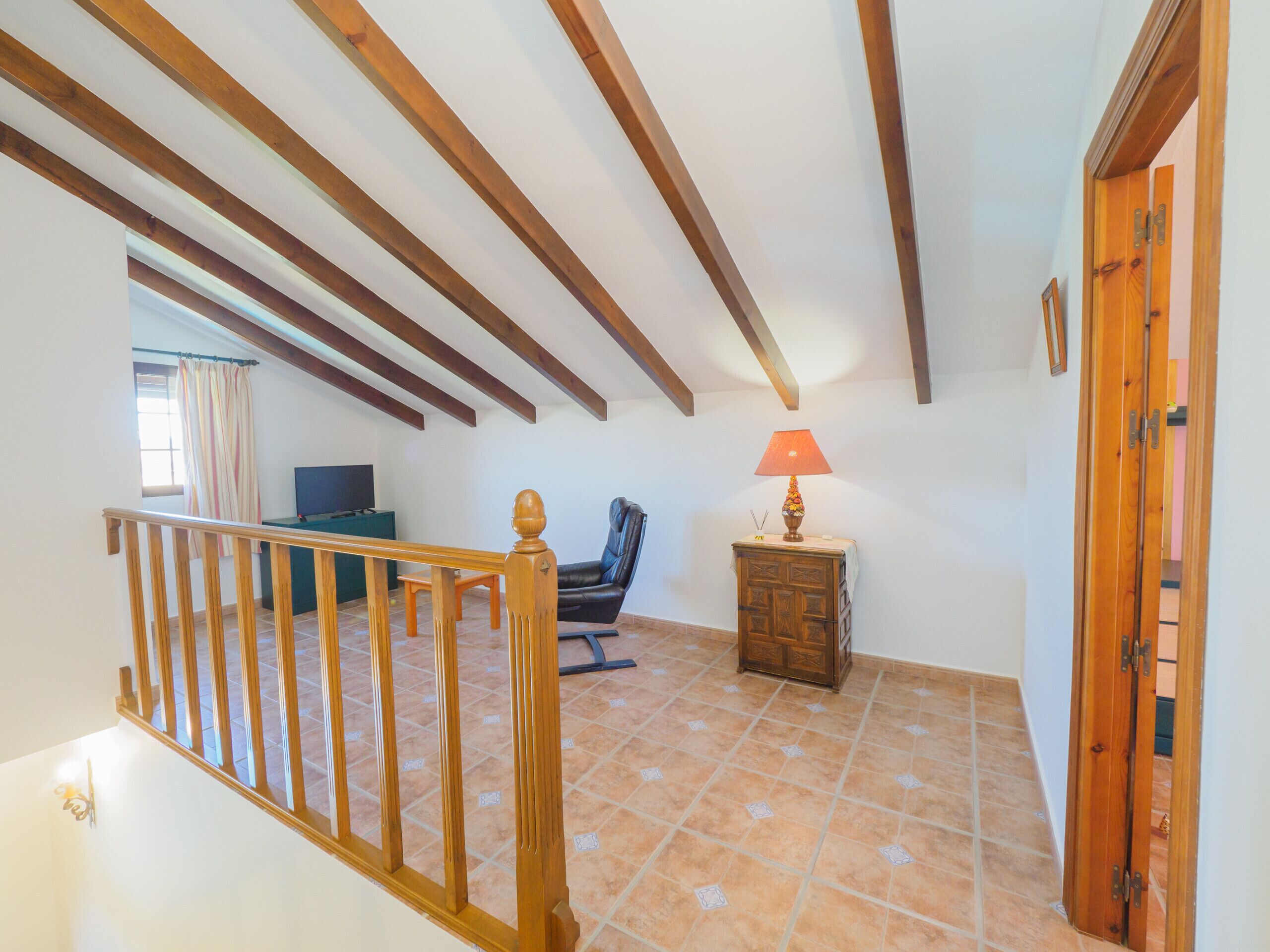 A spacious room with a high ceiling, exposed wooden beams, a tiled floor, a wooden staircase, a leather chair, a small wooden table with a lamp, and a television.