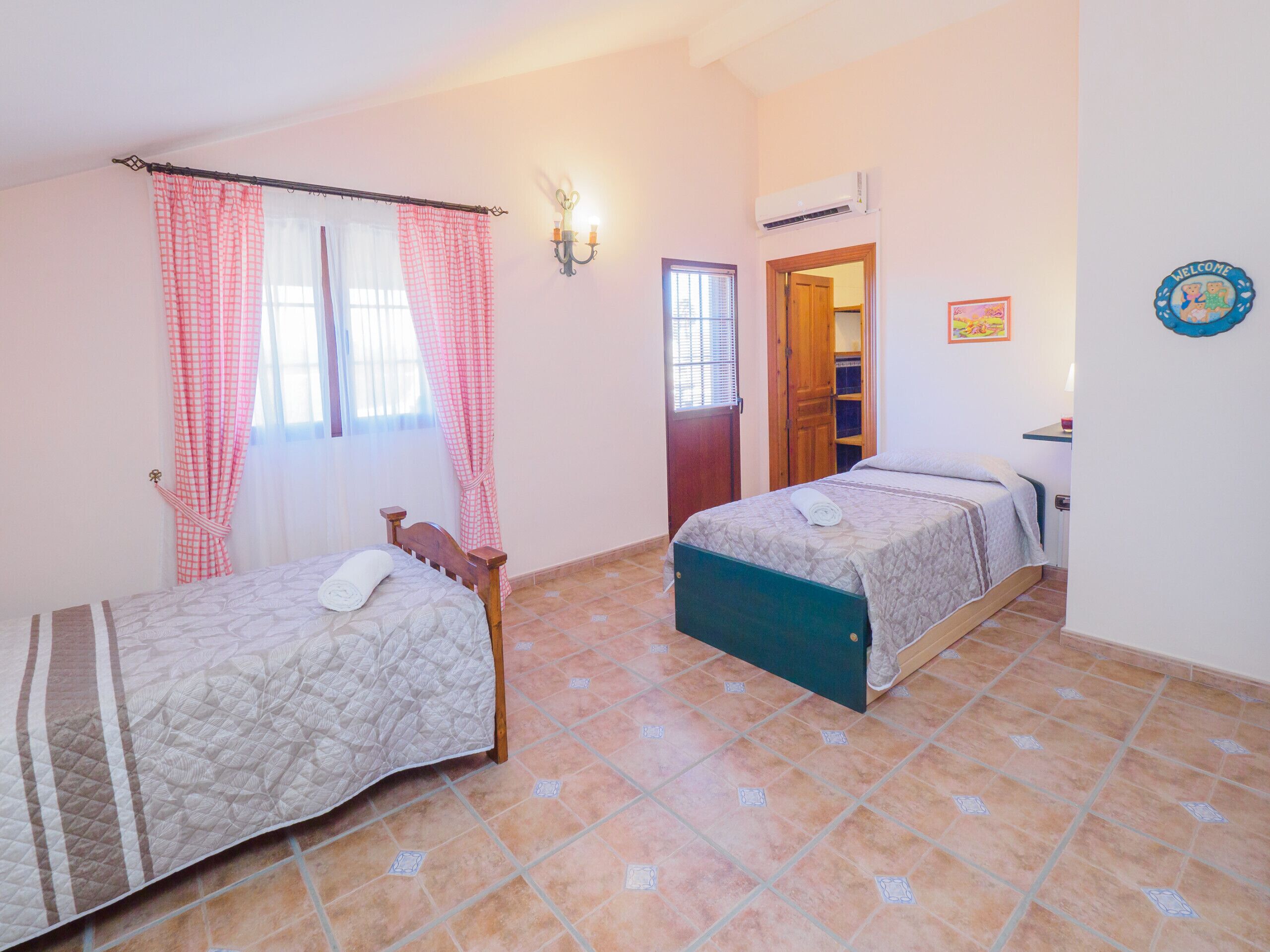 Une chambre avec deux lits, une fenêtre avec des rideaux, un climatiseur fixé au mur et une porte donnant sur une autre pièce.